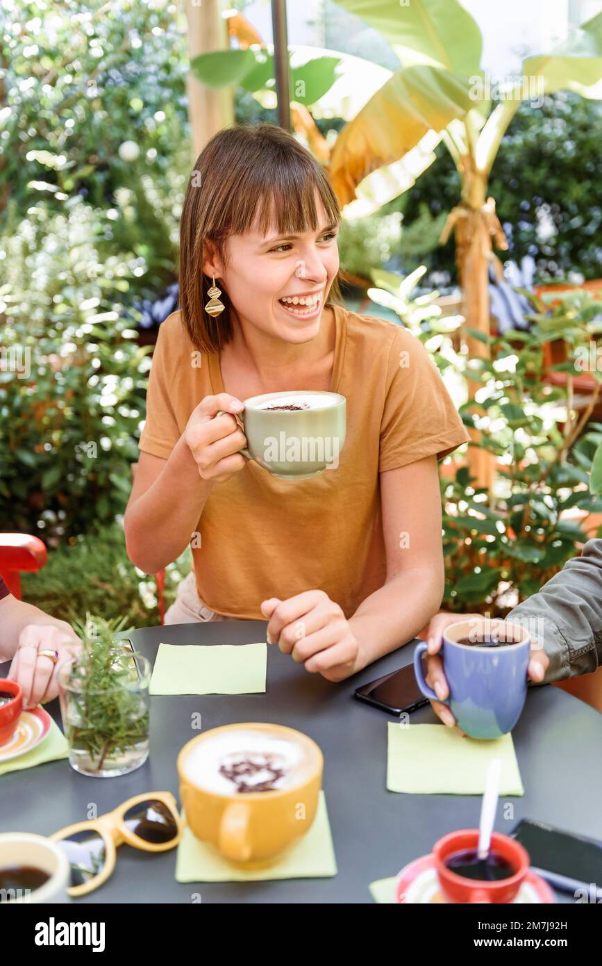 Der große Winkel einer fröhlichen jungen Frau mit einer Tasse Cappuccino, die über einen Witz lacht und wegschaut, während sie am Tisch sitzt, während sie sich mit Freunden in gar trifft Stockfoto
