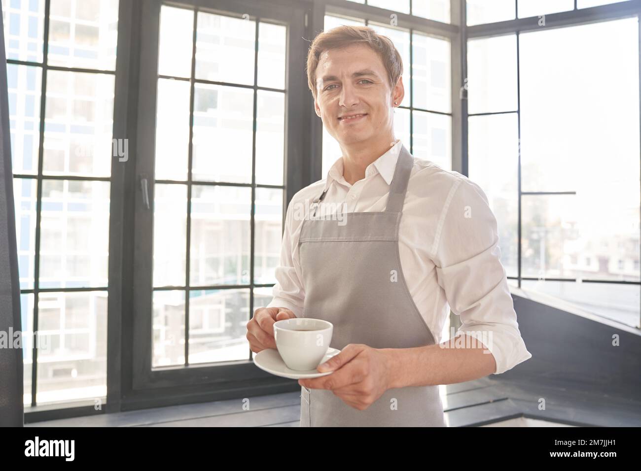 Ein Kellner in einer grauen Schürze hält eine Tasse Kaffee. Der Barista gibt eine Tasse heißen Kaffee in einem Café vor dem Hintergrund eines großen Fensters. Hochwertiges Foto Stockfoto
