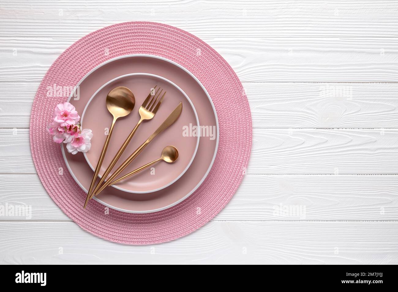 Rosafarbenes Geschirr und goldenes Besteck mit Blumen, Stille auf weißem Holztisch. Draufsicht. Geschirr und Utensilien zum Servieren und Essen. Moderner Tiger Stockfoto