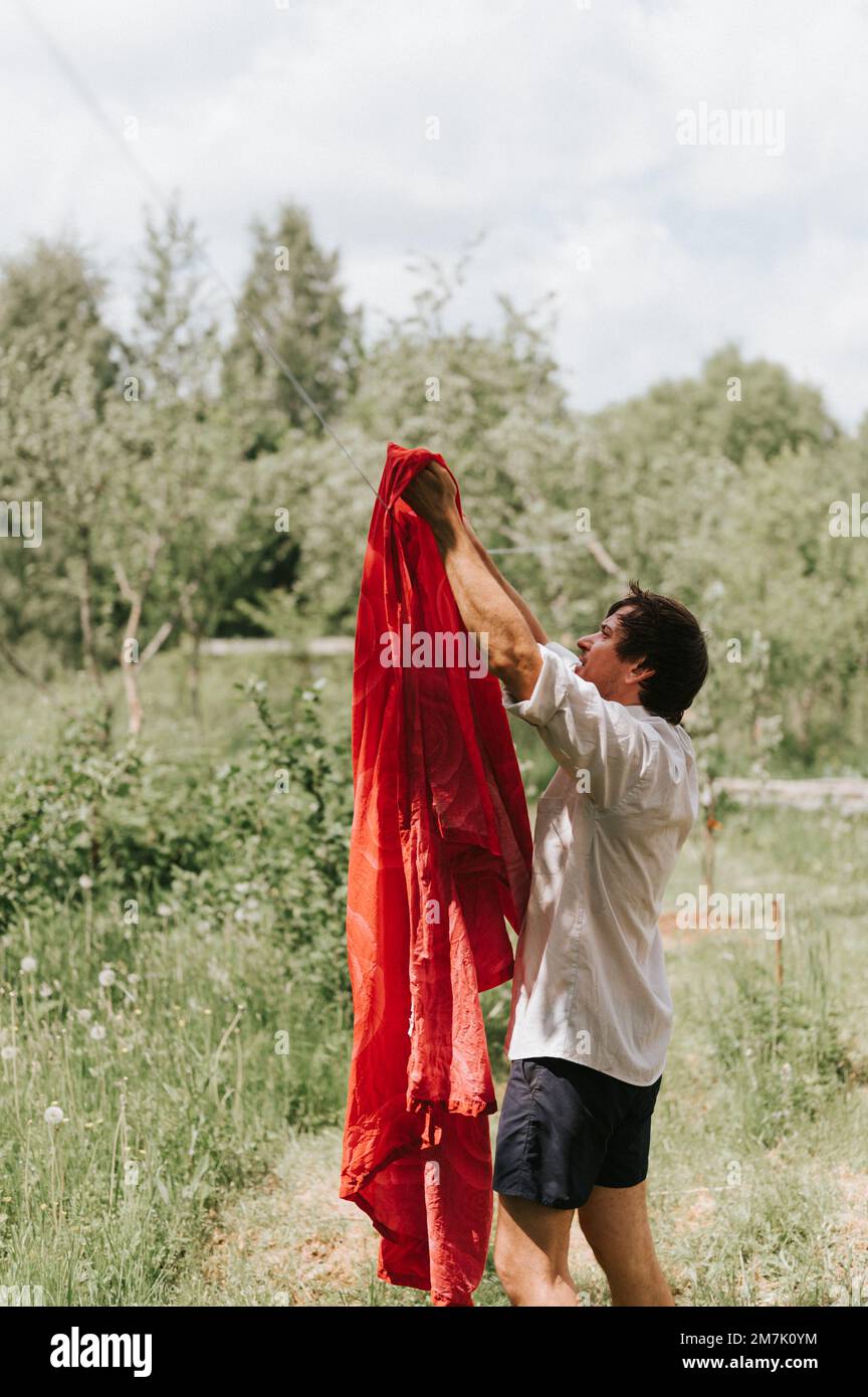 Ein aufrechter junger, reifer Hausangestellter macht Hausaufgaben und hängt die Wäsche zum Trocknen auf einer Wäscheleine auf der Straße im Innenhof des Dorfhaushaltes Stockfoto