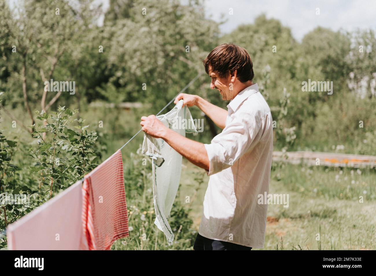 Ein aufrechter junger, reifer Hausangestellter macht Hausaufgaben und hängt die Wäsche zum Trocknen auf einer Wäscheleine auf der Straße im Innenhof des Dorfhaushaltes Stockfoto