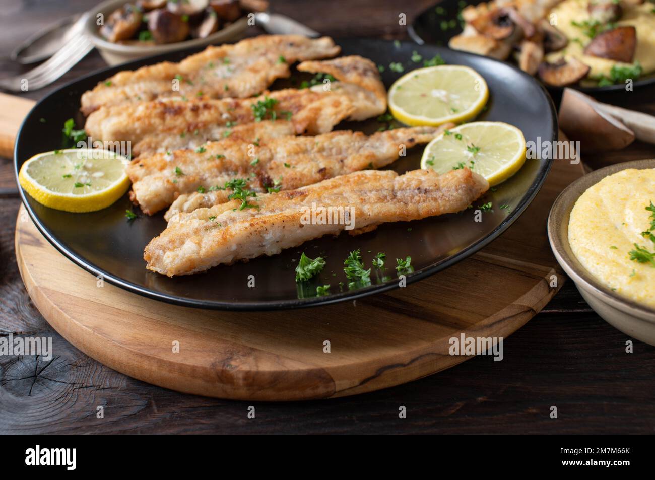 Gebratener Fisch in der Pfanne mit cremigen Polenta und Pilzen in Butter auf einem rustikalen Holztisch zum Abendessen oder Mittagessen Stockfoto