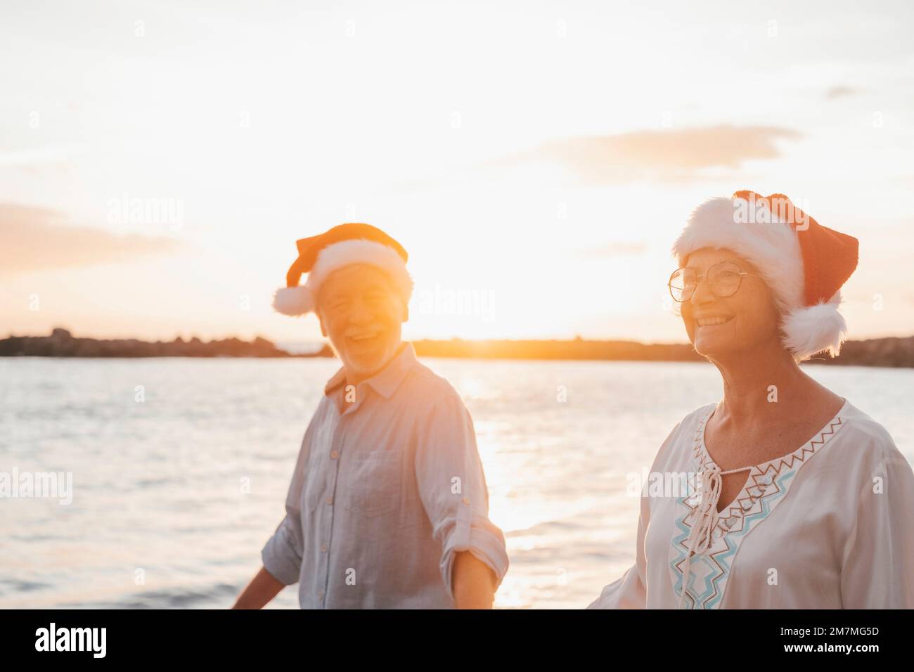 Ein altes, süßes Paar reifer Leute, die sich am Strand amüsieren und Spaß miteinander haben und an Feiertagen Weihnachtshüte tragen. Spaziergang am Strand mit Sonnenuntergang im Hintergrund im Winter. Stockfoto