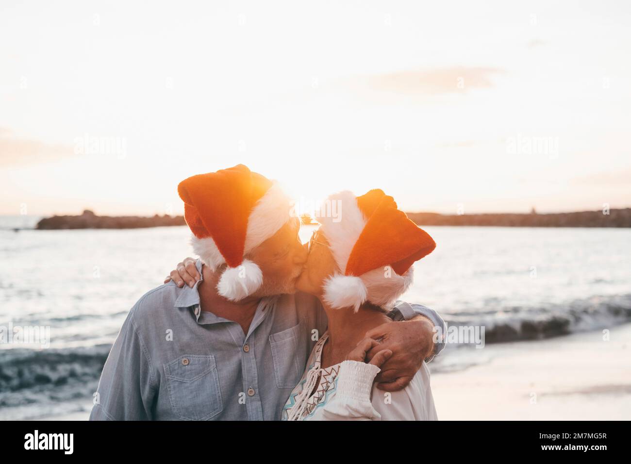 Ein altes, süßes Paar reifer Leute, die sich am Strand amüsieren und Spaß miteinander haben und an Feiertagen Weihnachtshüte tragen. Umarmt am Strand mit dem Sonnenuntergang im Hintergrund im Winter. Stockfoto