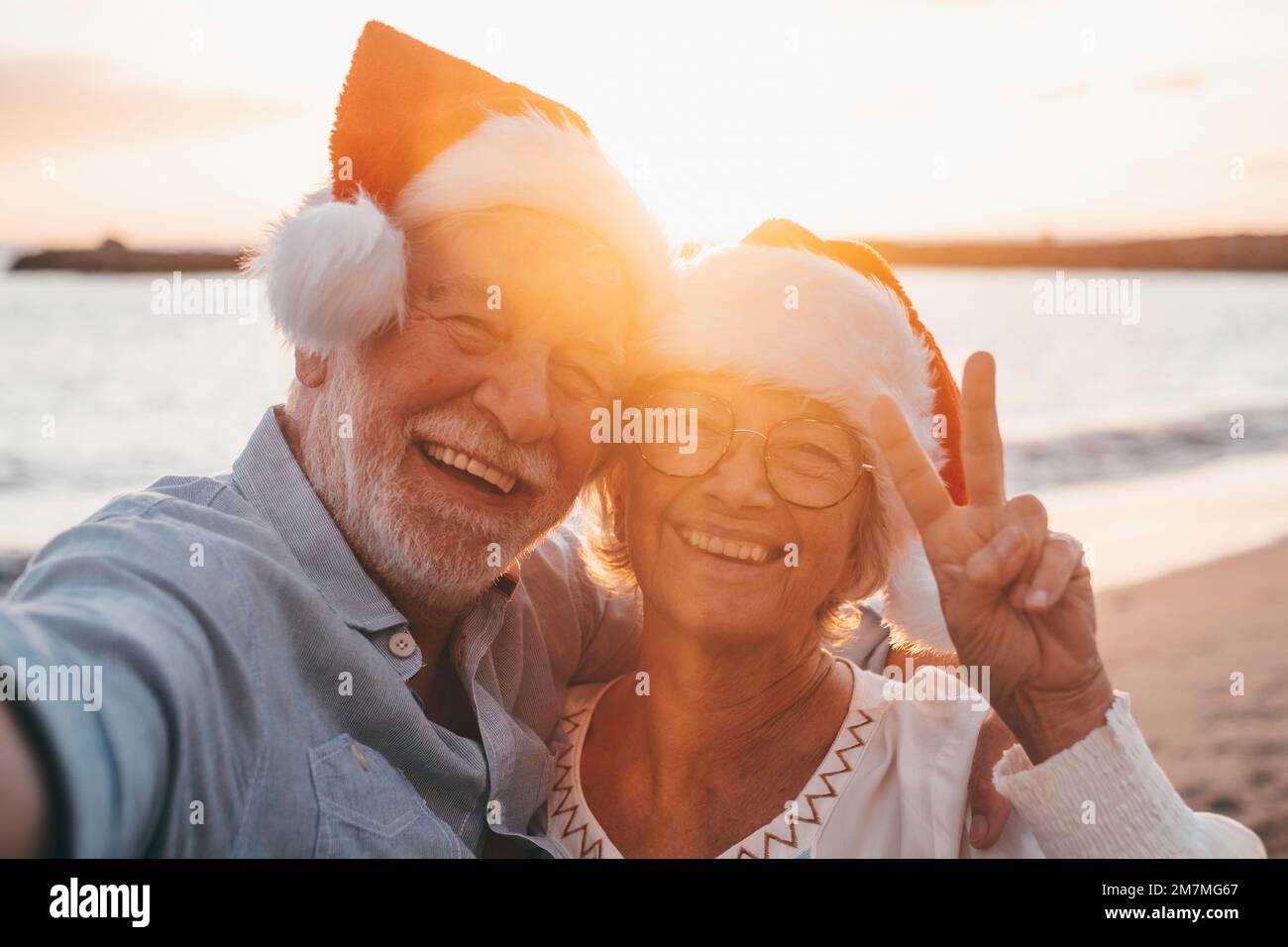 Porträt von zwei süßen alten Menschen, die Spaß haben und gemeinsam am Strand an weihnachtstagen am Strand mit Weihnachtshüten genießen. Ich schaue und halte eine Kamera, um Videos von Ferien aufzunehmen Stockfoto