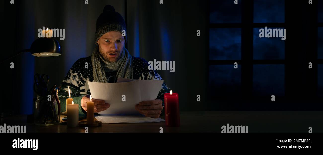 Ein schockierter Mann in Winterkleidung sieht sich die Haushaltsrechnungen an, während er in einem kalten Raum bei Kerzenschein sitzt. Energiekrise, Heizperiode Stockfoto