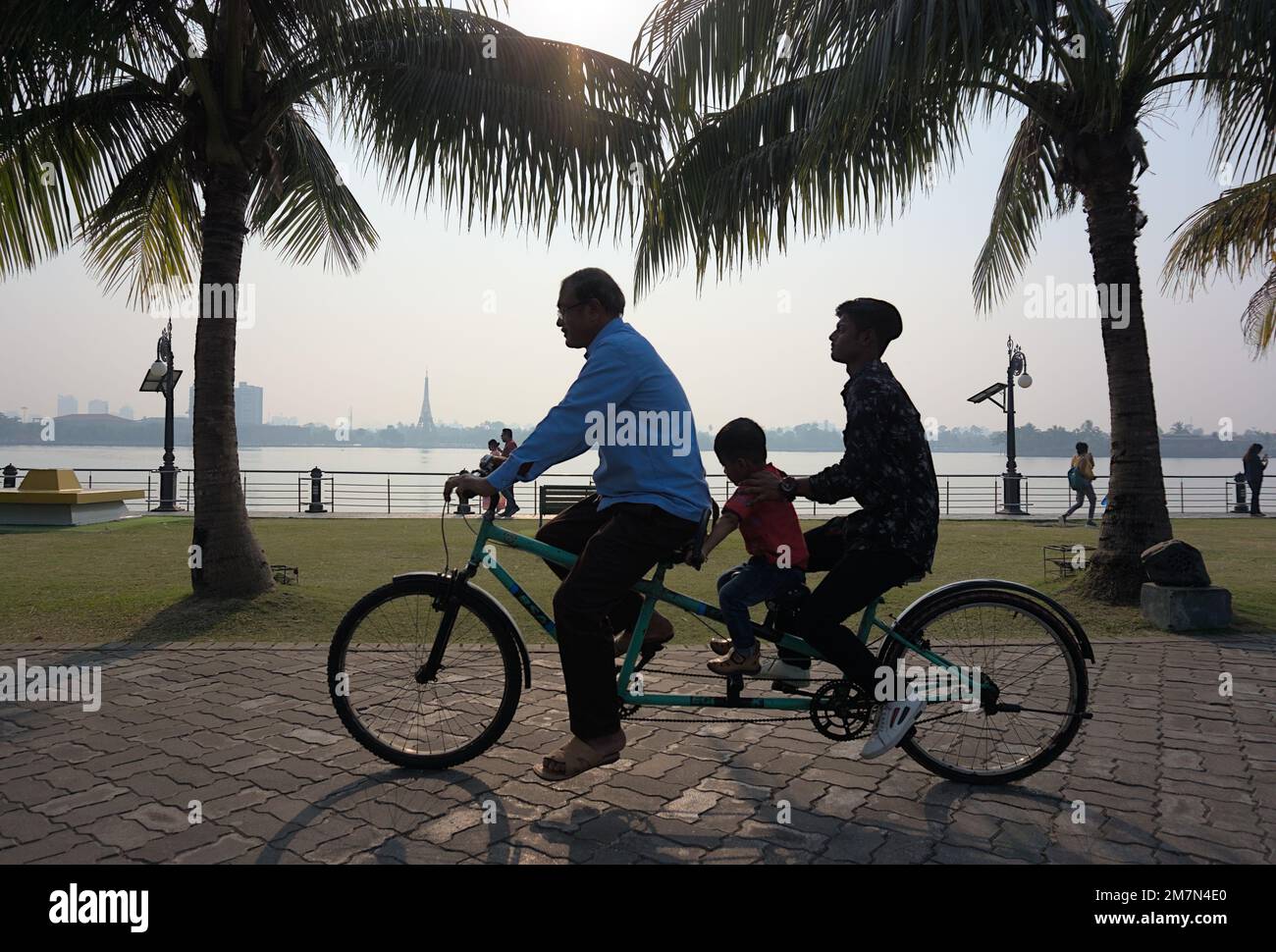 Drei Personen, die an einem sonnigen Tag an einem See ein Fahrrad namens Tandem Bike fahren. Drei Personen unterschiedlichen Alters, Erwachsene, Jugendliche, Kleinkinder. Stockfoto