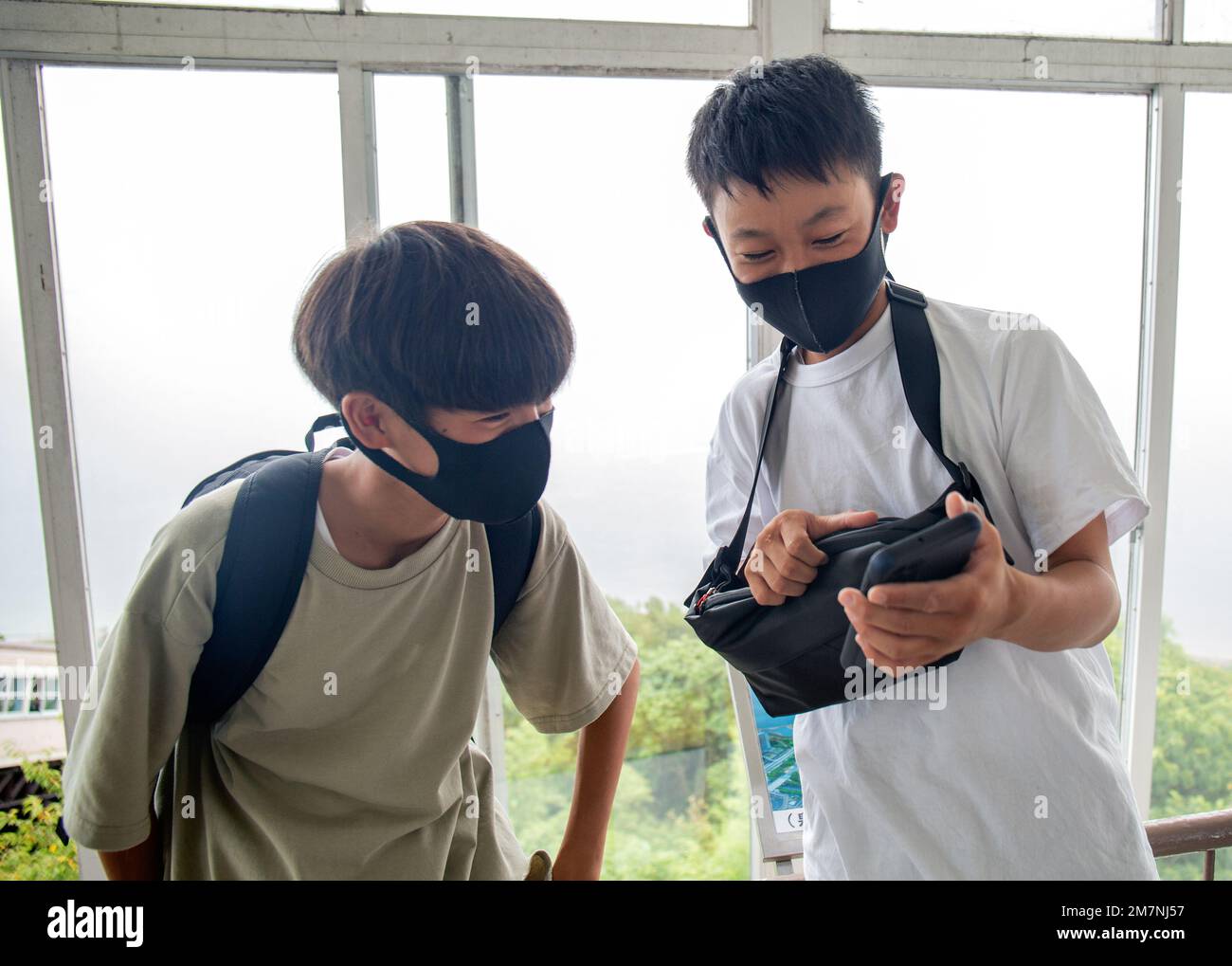 Zwei 13 Jahre alte japanische Jungen mit Gesichtsmasken, Freunde, die auf einen Handy-Bildschirm schauen. Stockfoto