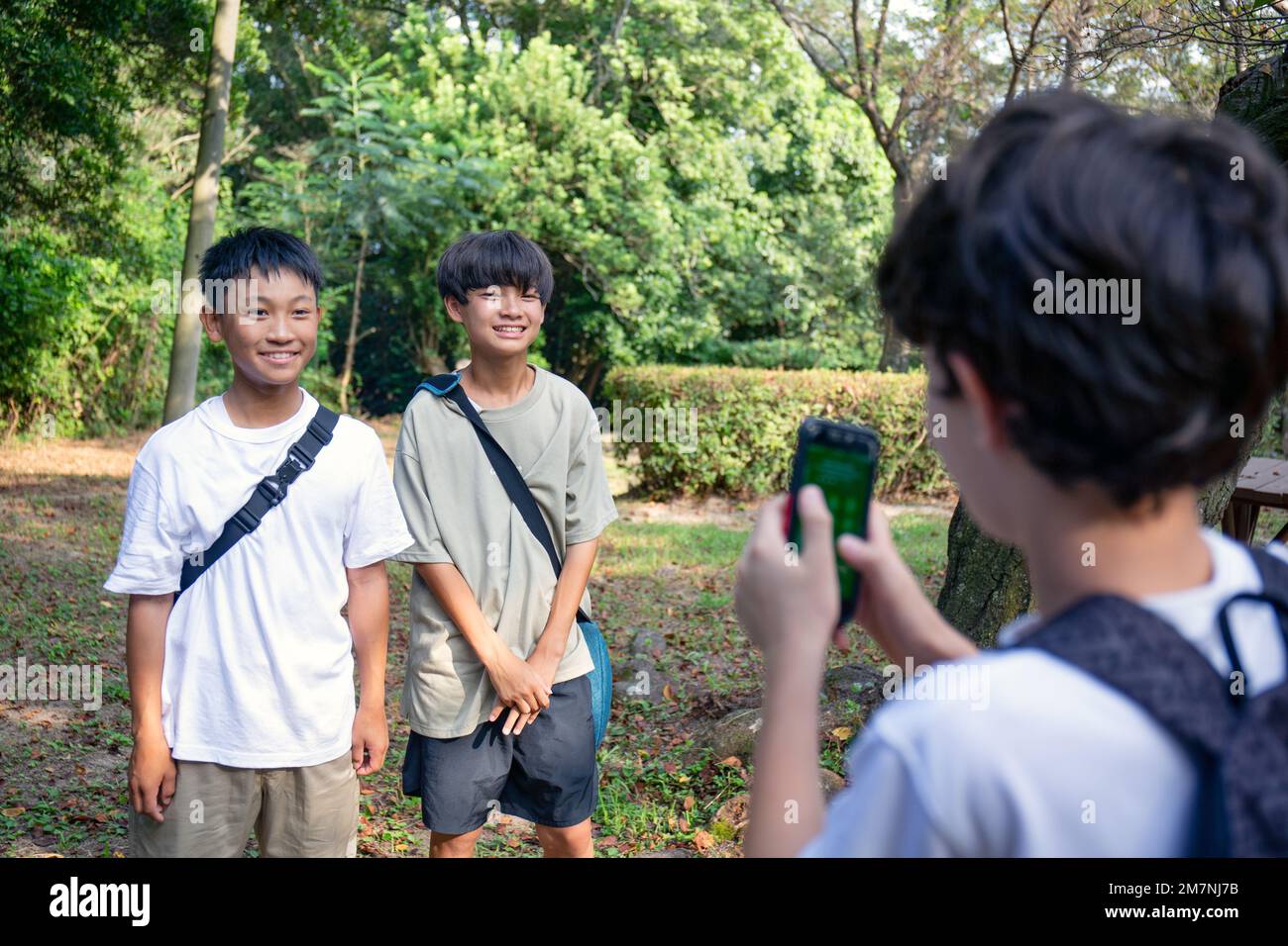 Ein Junge mit einem Handy, der im Sommer im Freien in einem Park ein Foto von zwei 13-jährigen Jungen nebeneinander macht. Stockfoto