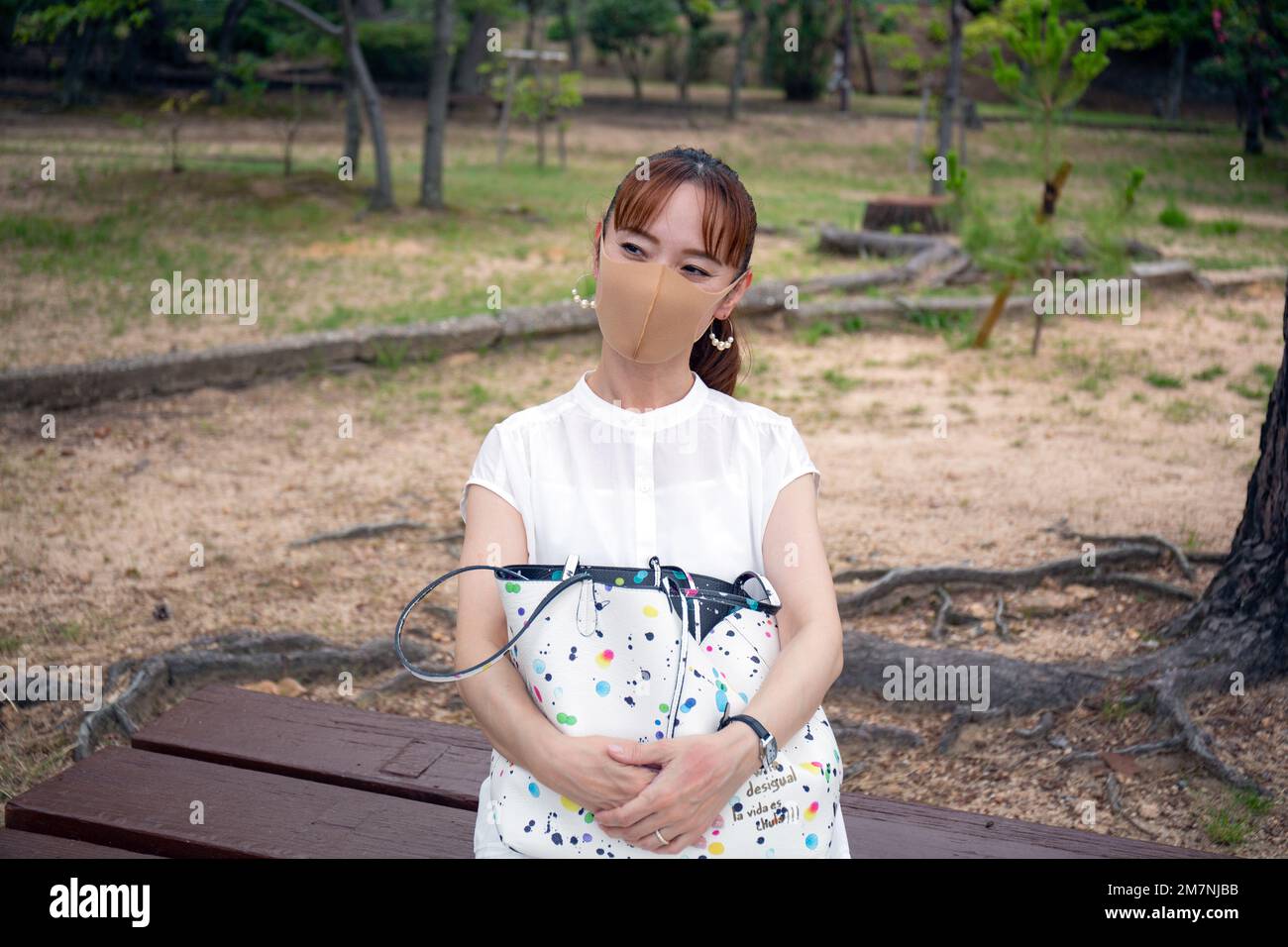 Eine reife japanische Frau draußen in einem Park, trägt eine Gesichtsmaske, hält eine Tasche in der Hand. Stockfoto