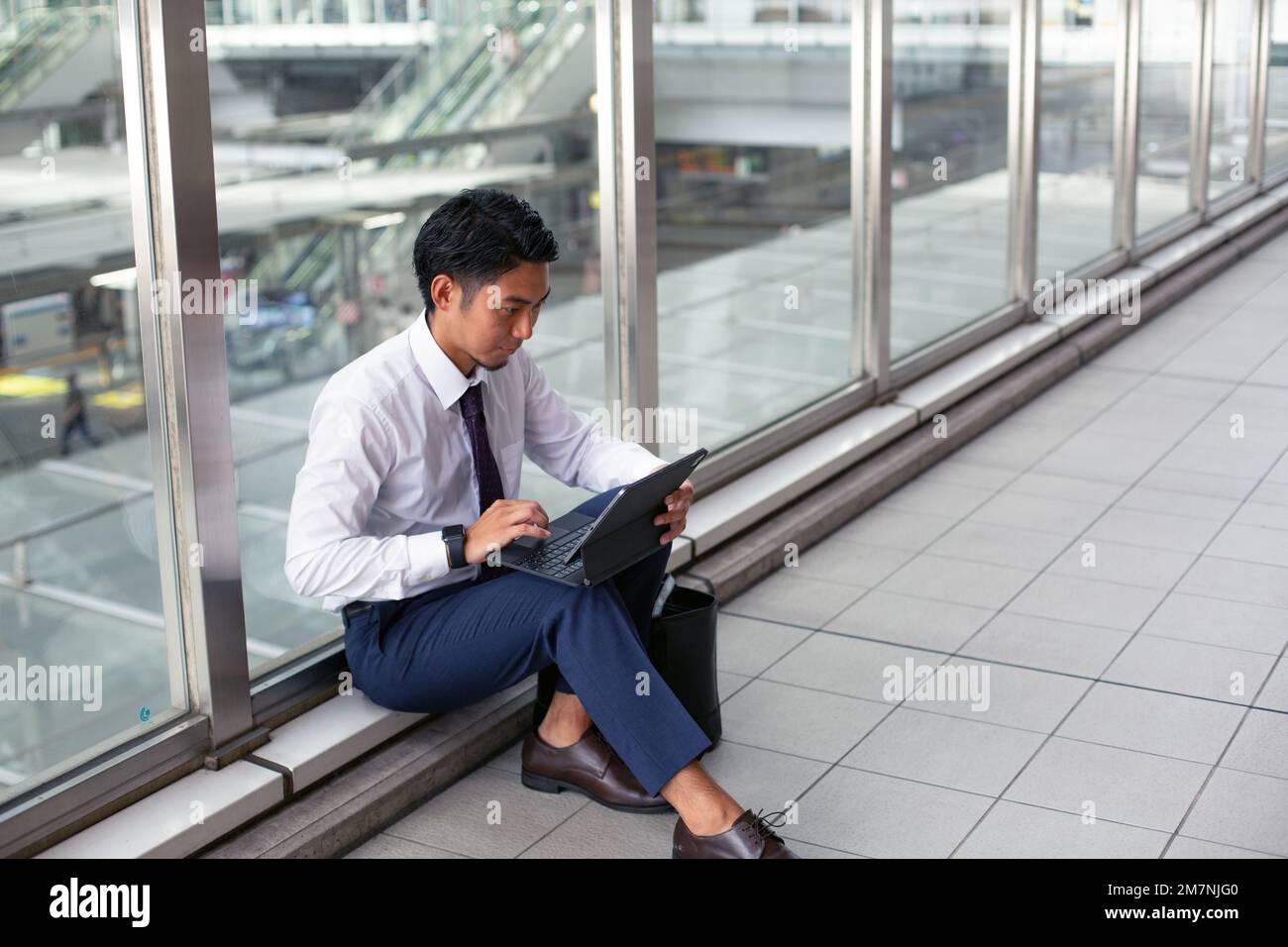 Ein junger Geschäftsmann in der Stadt, der unterwegs ist, auf einem Gehweg sitzt und seinen Laptop benutzt. Stockfoto