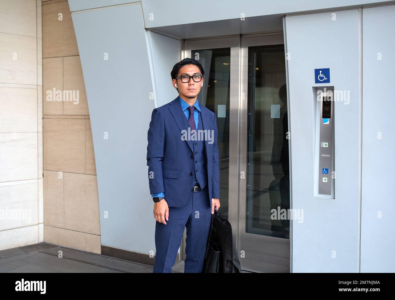Ein junger Geschäftsmann in der Stadt, der unterwegs ist, ein Mann, der an den Liftttüren steht, mit einer Laptop-Tasche und einem Anzug. Stockfoto