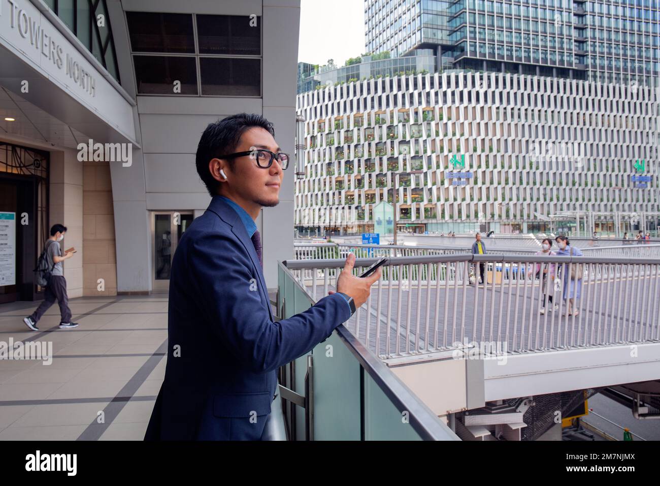 Ein junger Geschäftsmann in der Stadt, der unterwegs ist, ein Mann in einer blauen Jacke, der sein Handy in der Hand hält und Ohrstöpsel trägt. Stockfoto