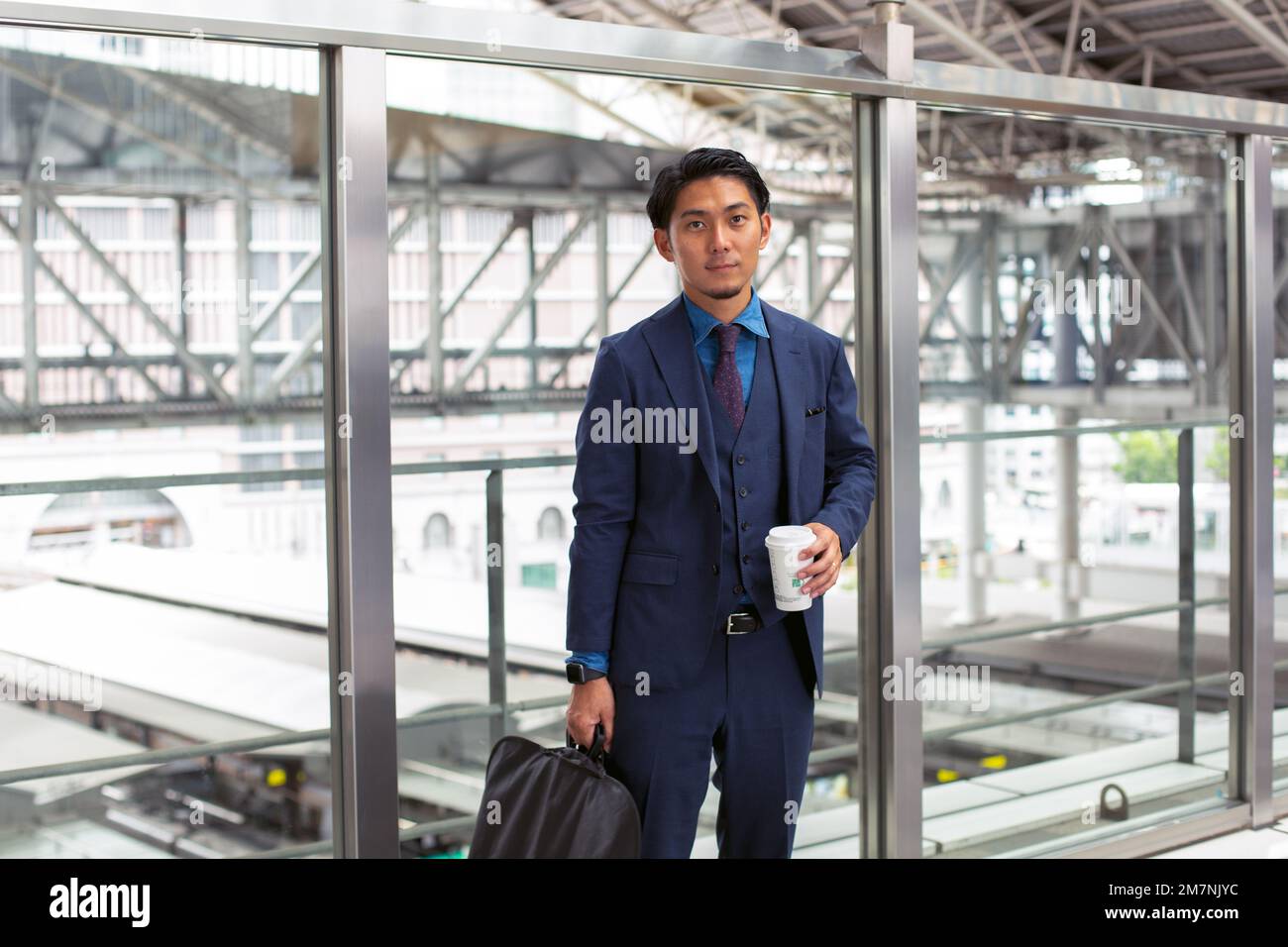 Ein junger Geschäftsmann in einem blauen Anzug, der in der Innenstadt unterwegs ist, mit einer Aktentasche und einer Tasse Kaffee. Stockfoto