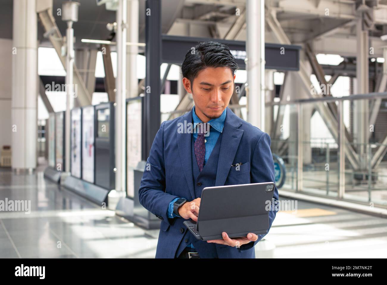 Ein junger Geschäftsmann in einem blauen Anzug, der unterwegs in einer Innenstadt ist, steht und benutzt sein digitales Tablet. Stockfoto