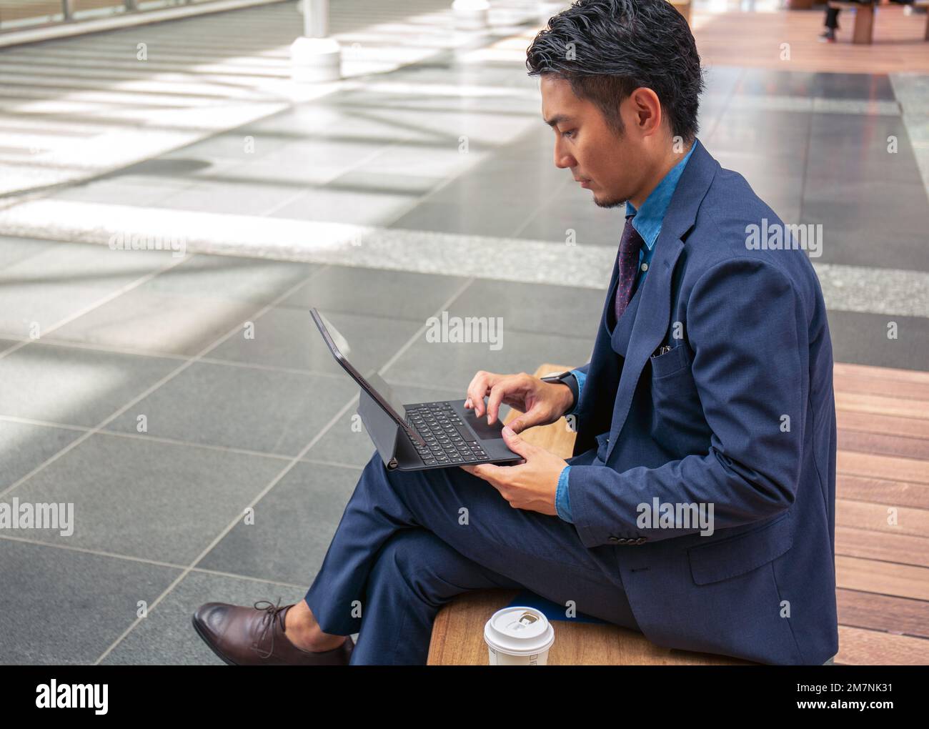 Ein junger Geschäftsmann in einem blauen Anzug, der unterwegs ist, in einer Innenstadt, sitzt auf einer Bank mit einem digitalen Tablet. Stockfoto