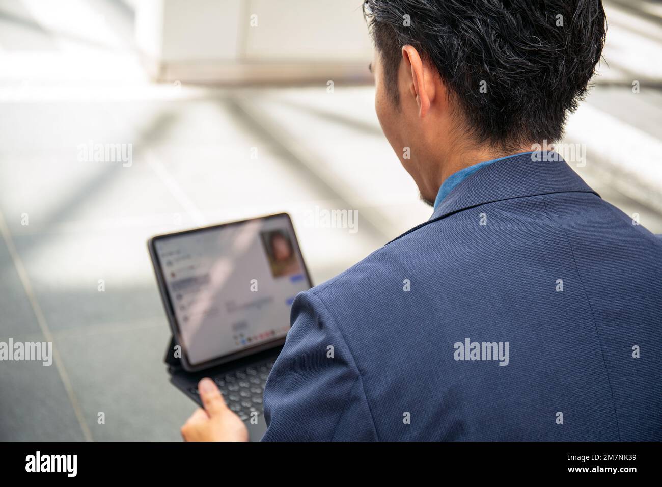 Ein junger Geschäftsmann in einem blauen Anzug, der unterwegs ist, in einer Innenstadt, sitzt auf einer Bank und benutzt ein digitales Tablet mit Bildschirm. Stockfoto