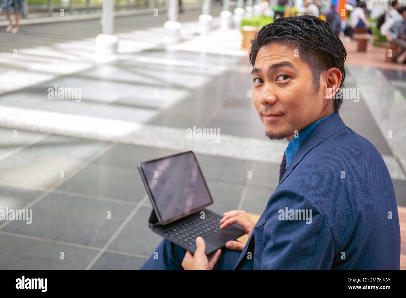 Ein junger Geschäftsmann in einem blauen Anzug in einer Stadtmitte, der mit einem digitalen Tablet auf einer Bank sitzt und über seine Schulter schaut. Stockfoto