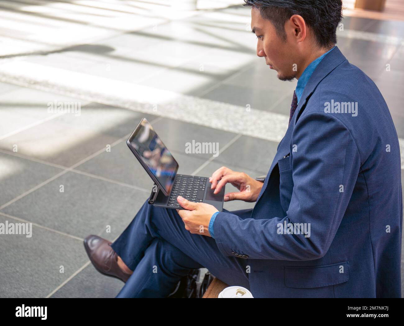 Ein junger Geschäftsmann in einem blauen Anzug, der unterwegs ist, in einer Innenstadt, sitzt auf einer Bank und benutzt ein digitales Tablet mit Bildschirm. Stockfoto