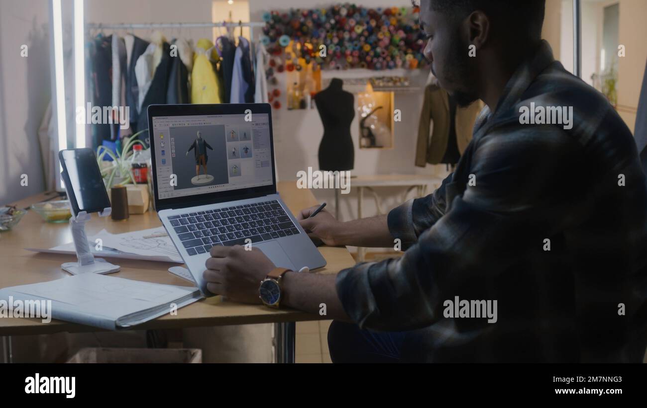 Afroamerikanischer Modedesigner entwickelt Bekleidungsdesign im 3D-Modellierungsprogramm auf einem Notebook mit digitalem Tablet und Eingabestift. Er arbeitet in der Atelier-Werkstatt. Mode und Technik im Geschäftskonzept. Stockfoto