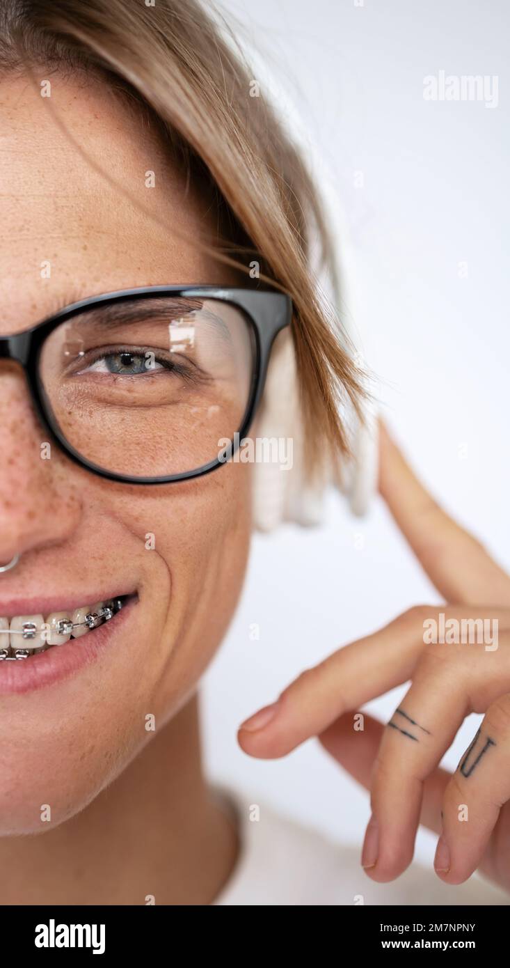 Verkürzte Porträtfrau mit Brille und Zahnbremsen, die Musik mit Kopfhörern hört und auf die Kamera schaut, wenn sie halb vor weißem Hintergrund steht. Stockfoto