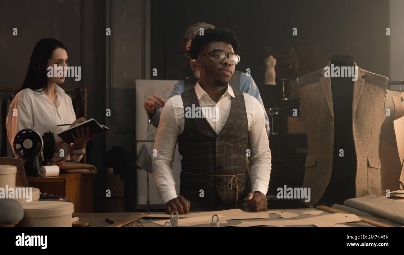 Männlicher Schneider misst afroamerikanische Klienten, diskutiert mit weiblicher Schneiderin Design von maßgeschneiderten Anzügen. Frau macht Notizen und schreibt die Maße auf. Konzept von Mode und Handwerk. Stockfoto