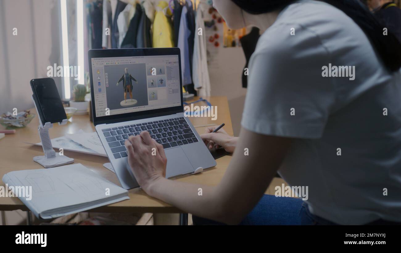 Weibliche Modedesignerin zeichnet zukünftige Kleidung in 3D-Modelliersoftware auf einem Laptop mit digitalem Tablet-Computer und Eingabestift. Sie arbeitet in einer Atelier-Werkstatt. Mode- und Technologiekonzept in der Wirtschaft. Stockfoto