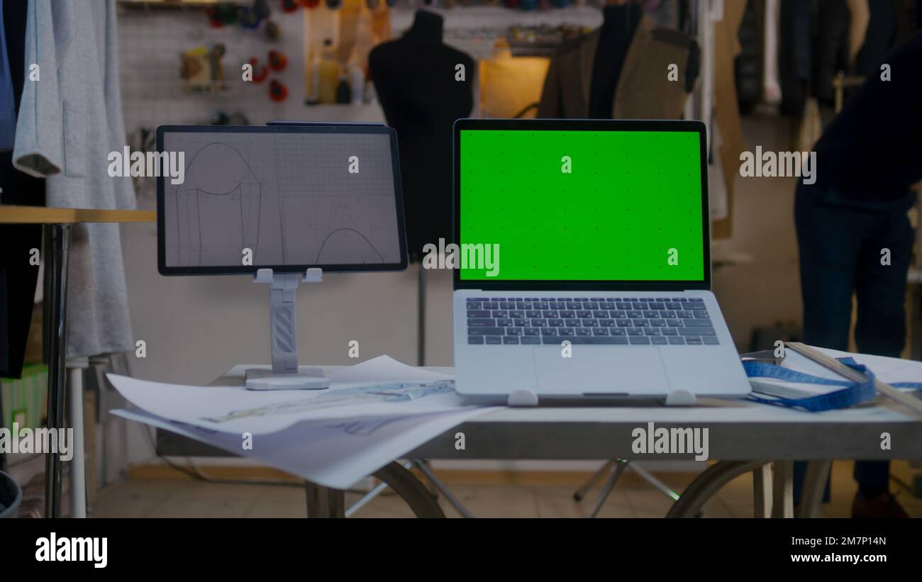 Dolly-Foto eines Laptops mit grünem Bildschirm und einem digitalen Tablet-Computer mit Skizze auf dem Tisch in einer Atelier-Werkstatt. Maßgeschneiderte und Schneiderarbeiten im Hintergrund. Mode- und Technologiekonzept in der Wirtschaft. Stockfoto