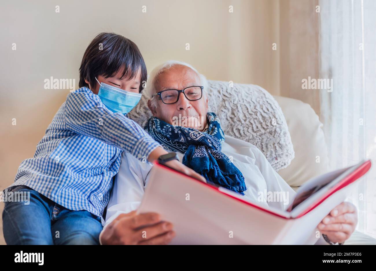Ein Großvater und sein Enkel lesen ein Buch und amüsieren sich im Wohnzimmer. Das Licht ist weich. Der Enkel trägt eine Maske. Stockfoto
