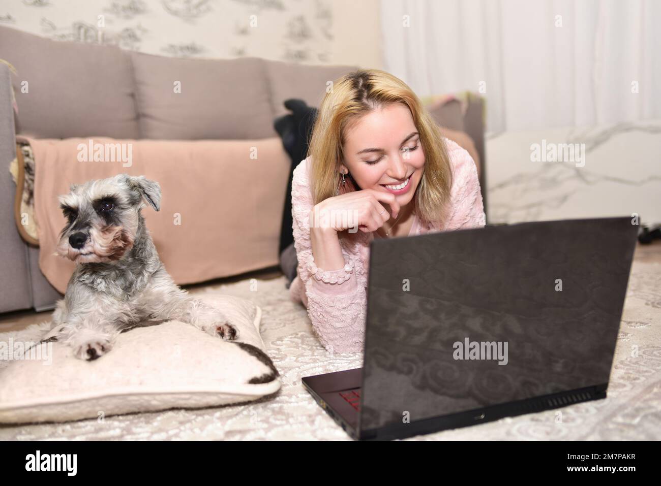 Eine junge blonde Frau lacht zu Hause mit einem Hund, während sie mit jemandem über ein Videogespräch auf einem Laptop chattet. Stockfoto
