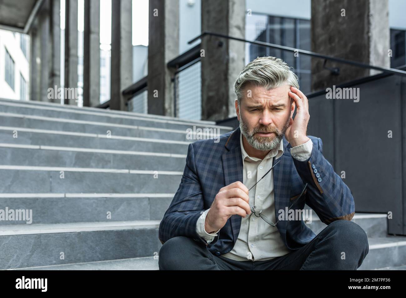 Grauhaariger Geschäftsmann, der traurig auf der Treppe vor dem Bürogebäude saß, deprimierter Chef im Business-Anzug, verärgert und enttäuscht von den Ergebnissen. Stockfoto