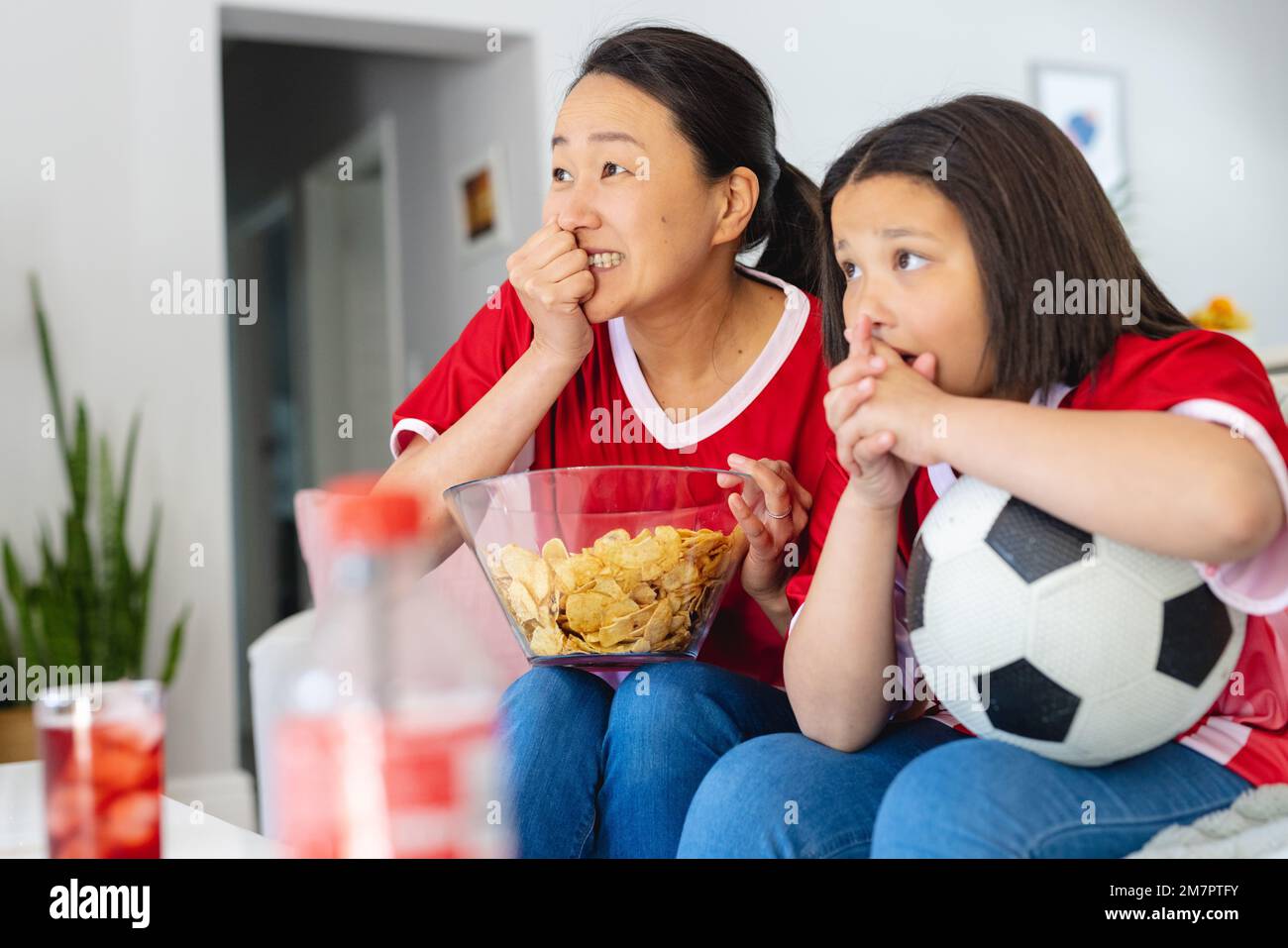 Asiatische Mutter und Tochter in Fußballtrikots, die Fußballspiele anschauen, Chips unterstützen und essen Stockfoto