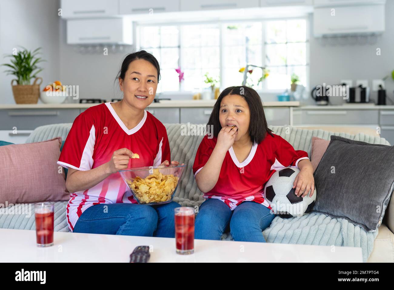 Asiatische Mutter und Tochter in Fußballtrikots, die Fußballspiele anschauen, Chips unterstützen und essen Stockfoto