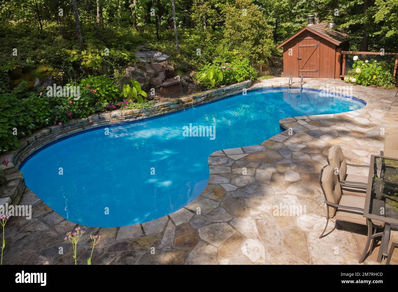 Im Erdgeschoss gelegener Swimmingpool mit Wasserfall und Steinplatten-Terrasse im landschaftlich gestalteten Hinterhof im Sommer. Stockfoto