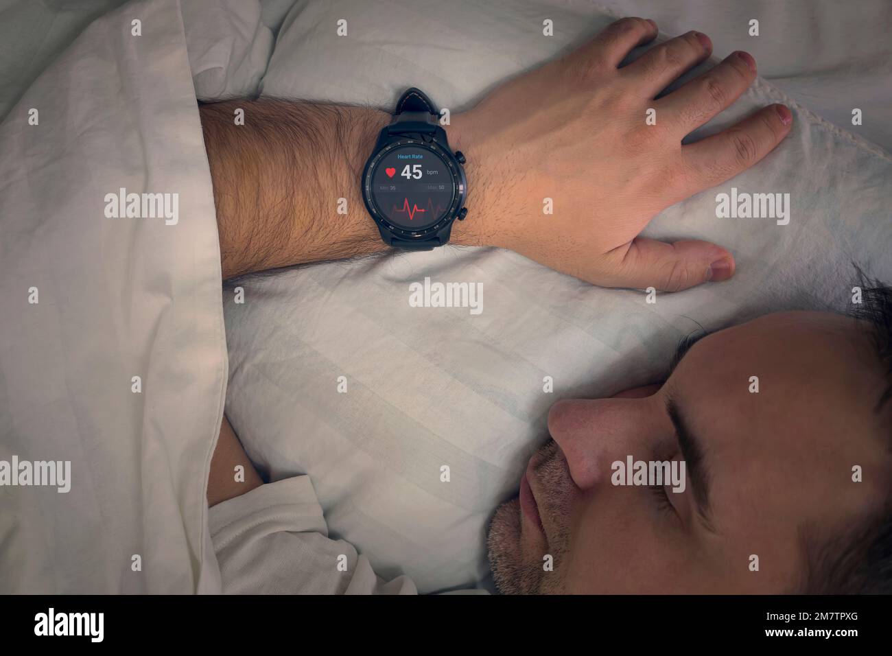 Fitness Activity Tracker mit Heartbeat Rate on man's Hand Over Bed. Ein Mann schläft auf einem Bett mit einer Herzfrequenz-Kontrolle in seinem Schlaf. Intelligente Uhren messen Stockfoto