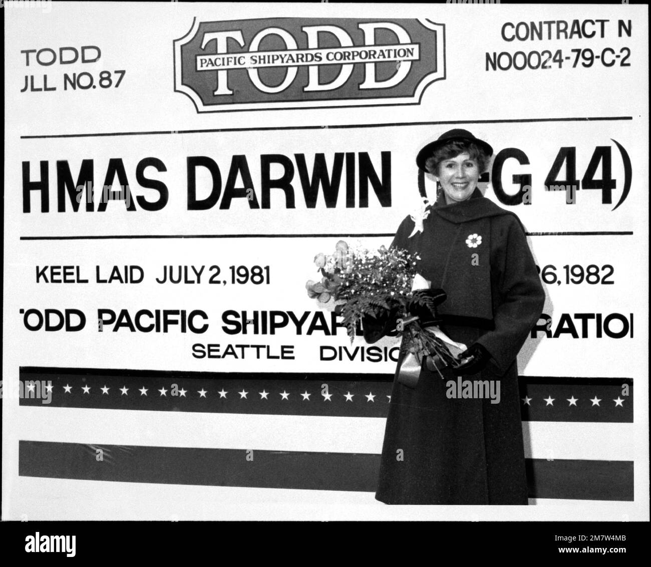 Sponsorin Mrs. E.E. Johnson hält Blumen vor dem Schild der Todd Pacific Shipyard Corp. Für die Australian Guided Missile Freggate HMAS DARWIN (FFG-44), bevor die Startzeremonien beginnen. Basis: Seattle Staat: Washington (WA) Land: Vereinigte Staaten von Amerika (USA) Stockfoto