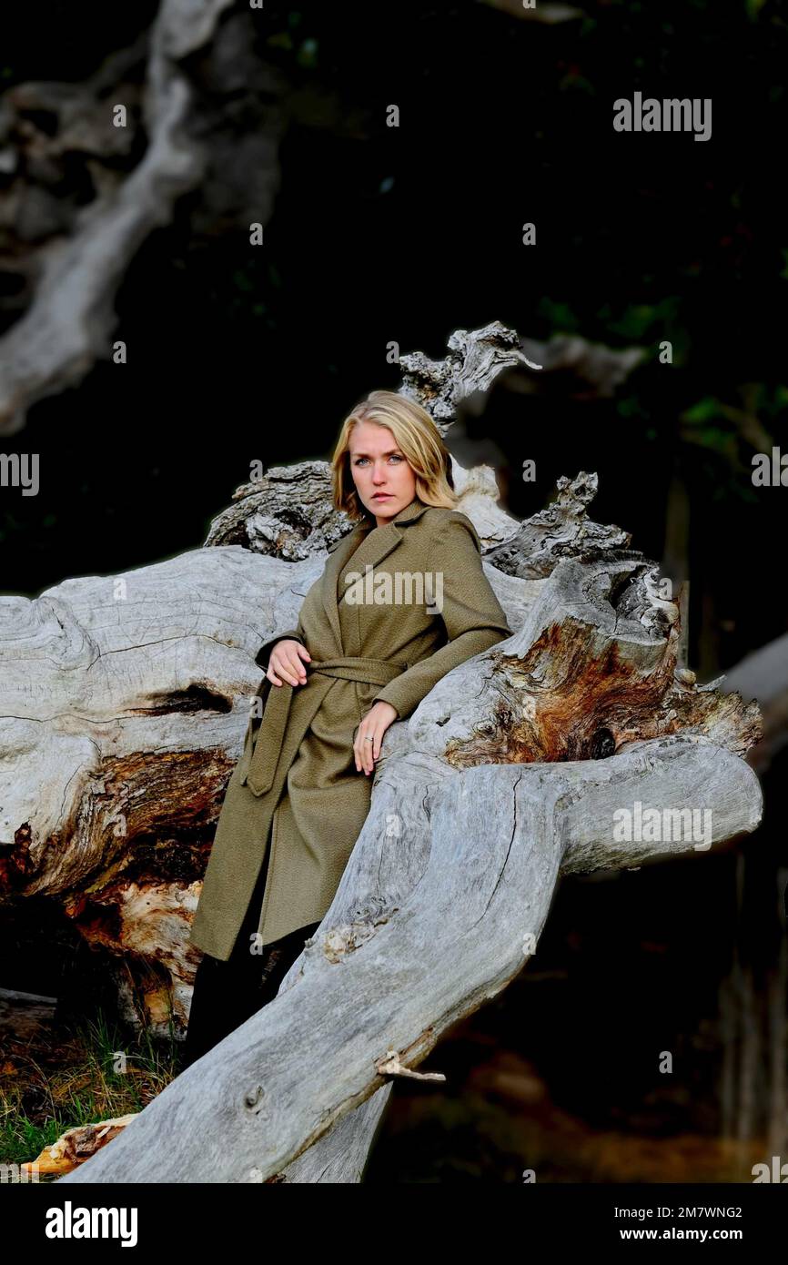 Eine junge blonde Frau im grünen Mantel, die sich gegen einen toten Baumstamm lehnt. Stockfoto