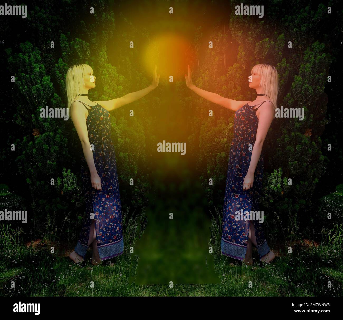 Junge blonde Frau in blauem Kleid mit Spiegelbild und mit glühender Kugel, die zwischen ihrer erhobenen Hand schwebt, großer Busch hinter ihr. Stockfoto