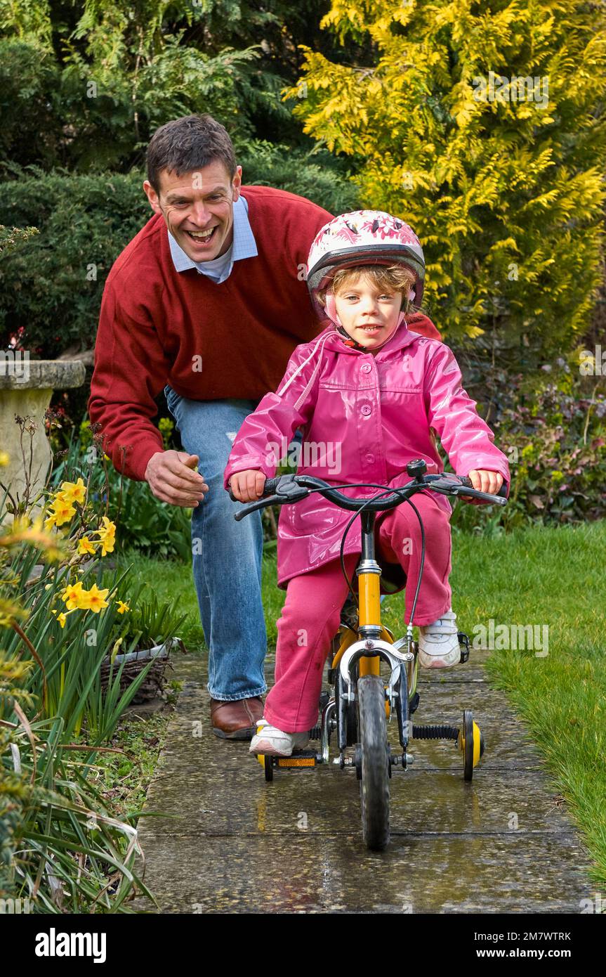 Ein Mann, der einem jungen Mädchen mit Stabilisatoren auf dem Weg in einem Garten hilft, ihr gelbes Fahrrad zu fahren Stockfoto