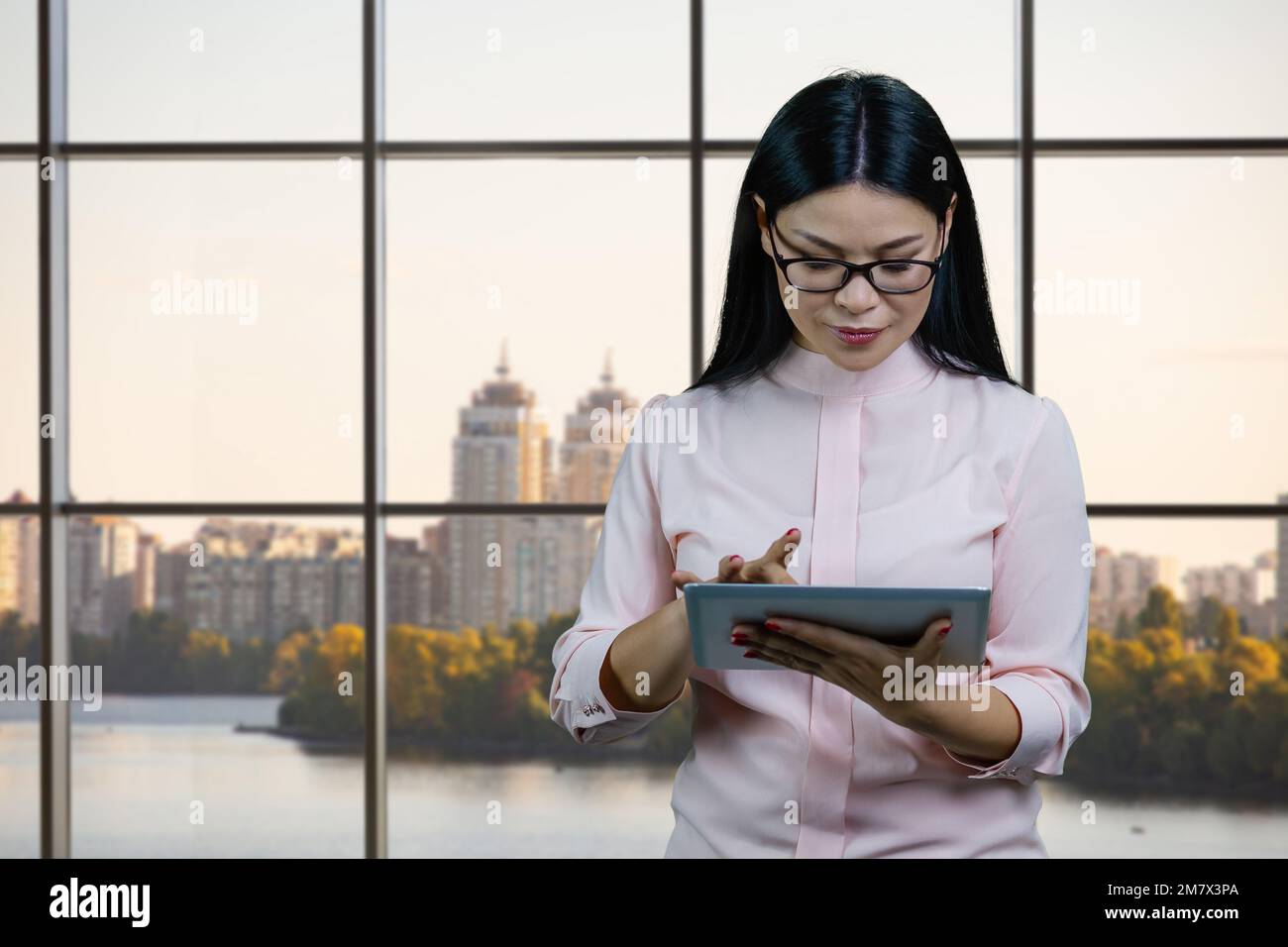 Eine junge asiatische Geschäftsfrau mit Brille verwendet im Büro einen Tablet-pc. Fenster und Hintergrund in Großstädten. Stockfoto
