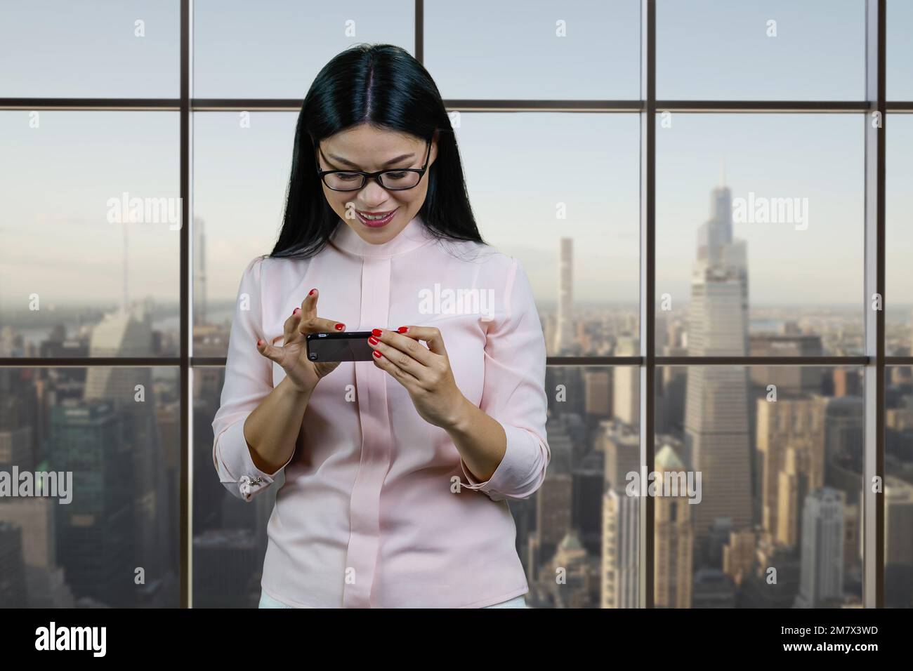Eine junge asiatische Geschäftsfrau zoomt etwas auf ihrem Smartphone ein. Fenster mit Stadtbild-Hintergrund. Stockfoto