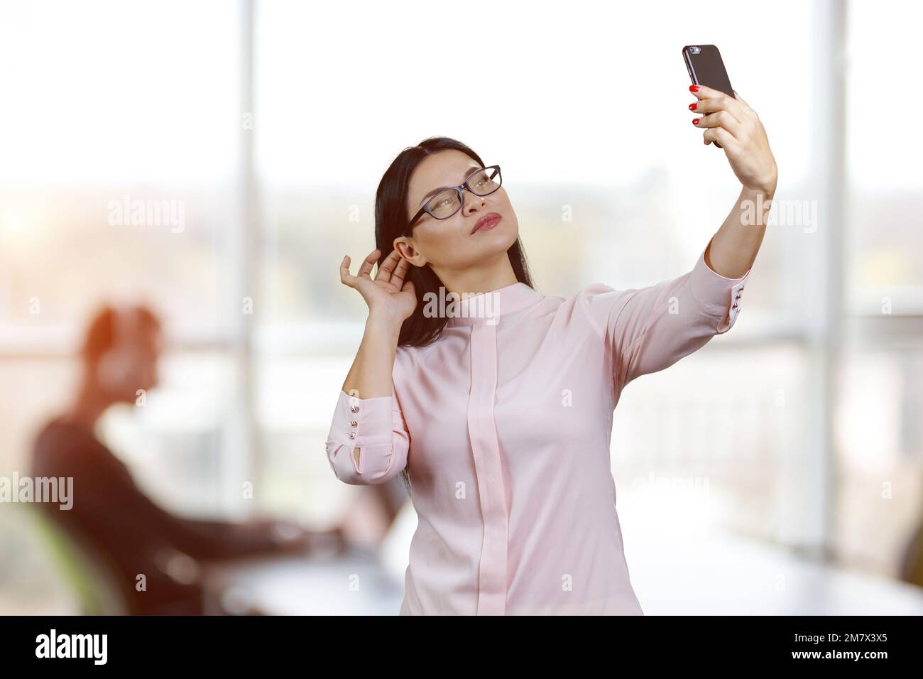 Junge asiatische Geschäftsfrau mit Brille macht ein Selfie. Bürofenster im Hintergrund. Stockfoto