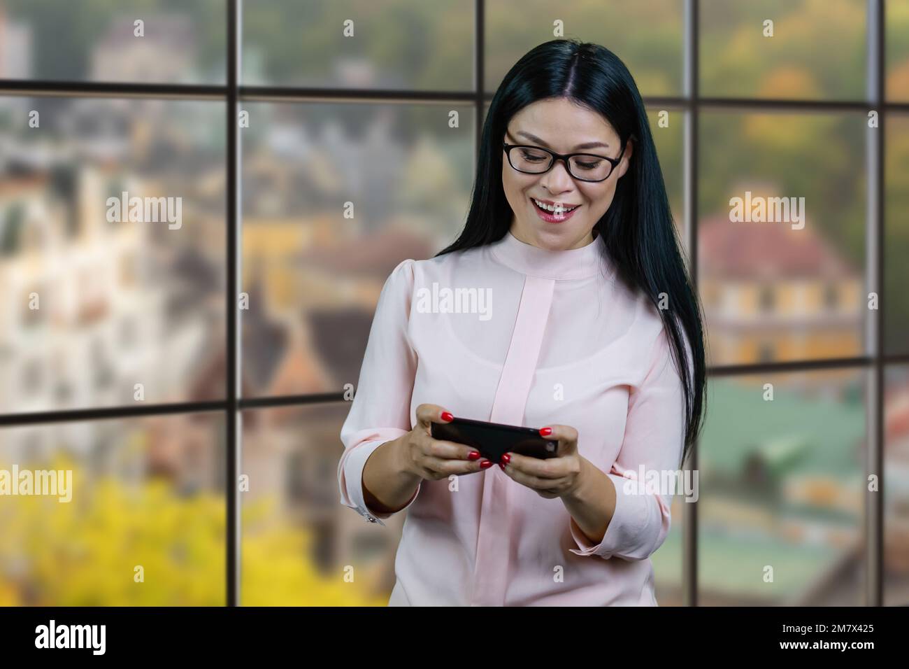 Glückliche asiatische Geschäftsfrau, die Videospiele auf ihrem Smartphone spielt. Karierte Fenster mit unscharfem Hintergrund. Stockfoto