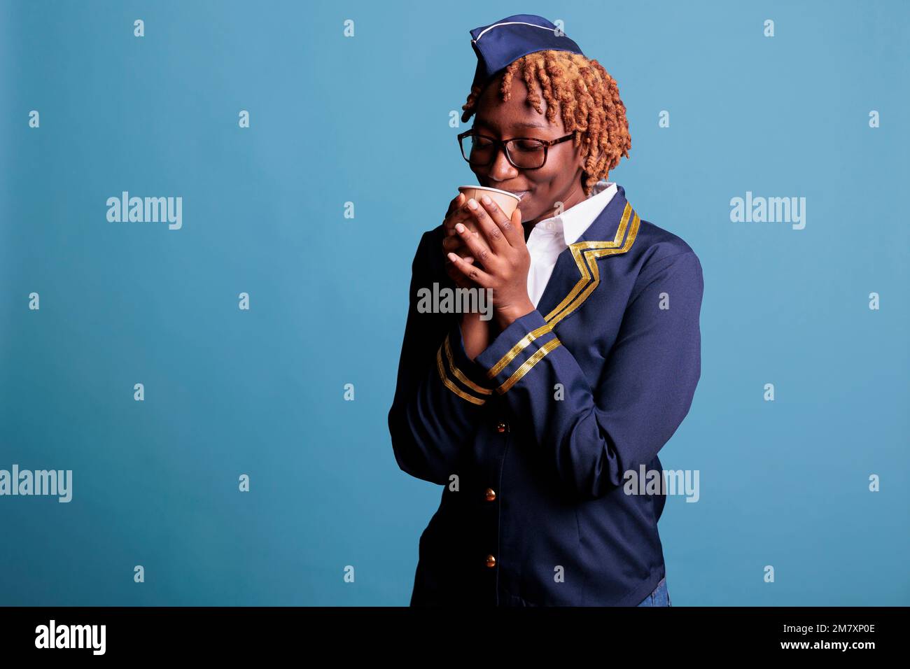 afroamerikanische Flugbegleiterin genießt eine Tasse heißen Kaffee, bevor die Passagiere an Bord gehen. Stewardess trägt Arbeitskleidung im Studio, aufgenommen vor blauem Hintergrund. Stockfoto