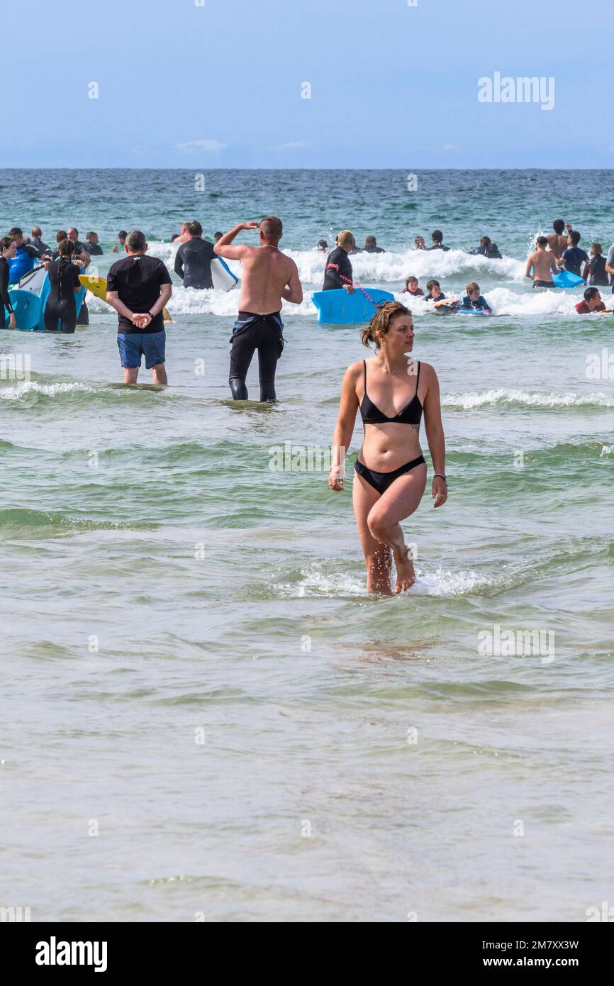 Eine Frau in einem Bikini, die auf einem geschäftigen Fistral Beach in Newquay in Cornwall in Großbritannien aus dem Meer spaziert. Stockfoto