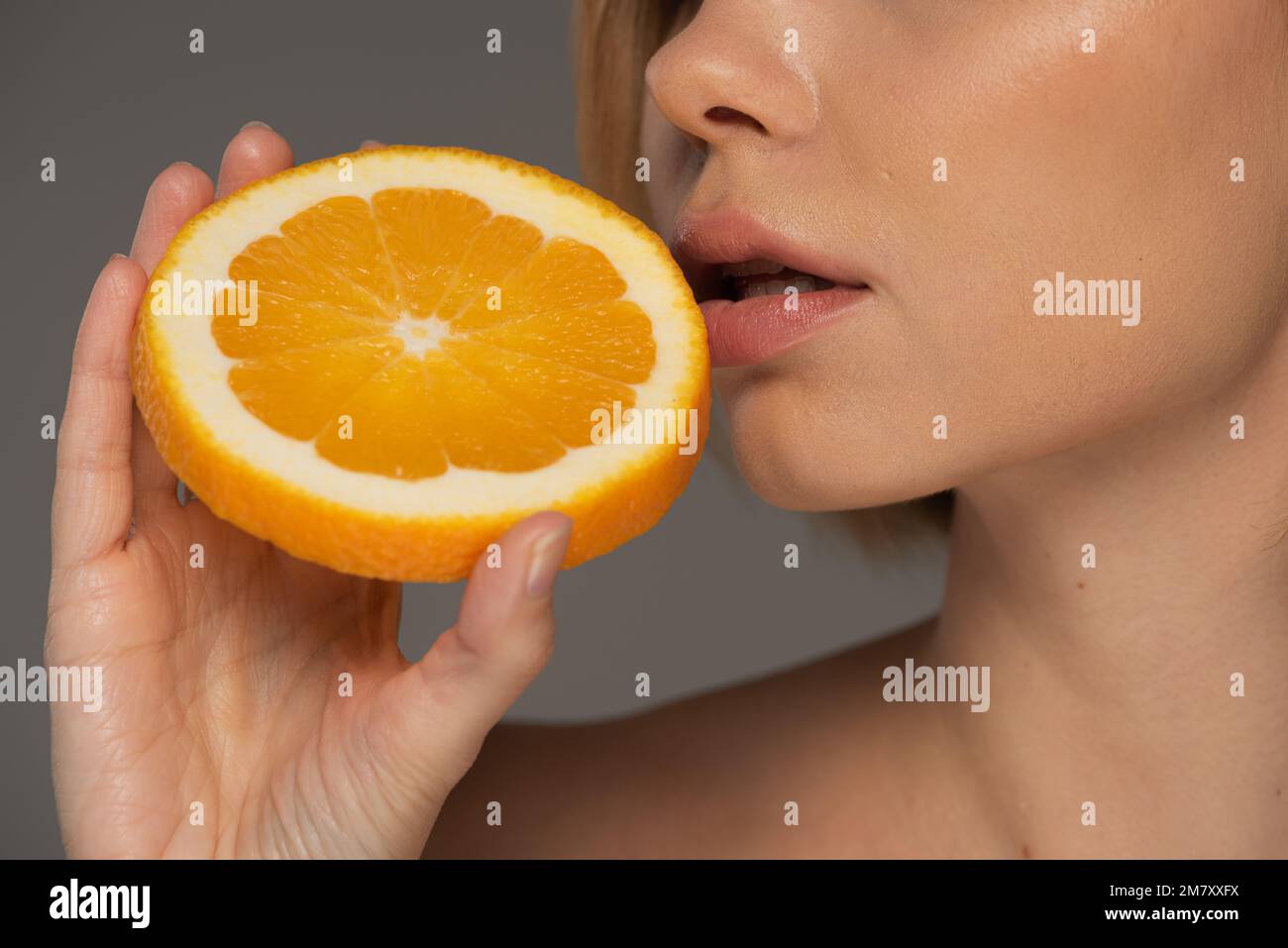 Zugeschnittene Ansicht einer jungen Frau, die eine saftige orangefarbene Hälfte in der Nähe weicher Lippen hält, isoliert auf einem grauen Stockbild Stockfoto