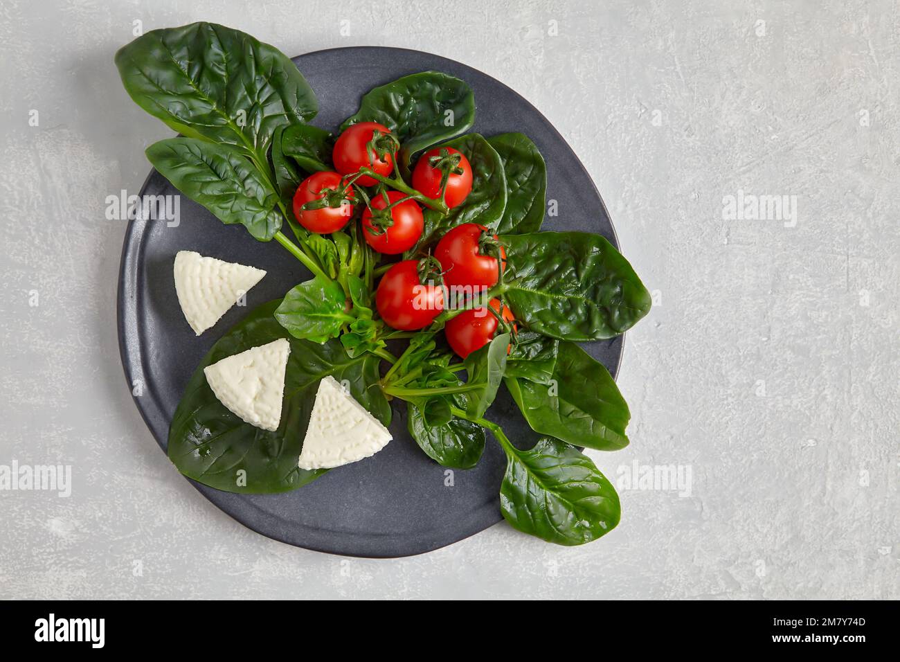 Rote Kirschtomaten auf einem Zweig, grüne Spinatblätter und Mozzarella-Quark auf einer dunklen Platte auf einem hellen Betontisch Stockfoto