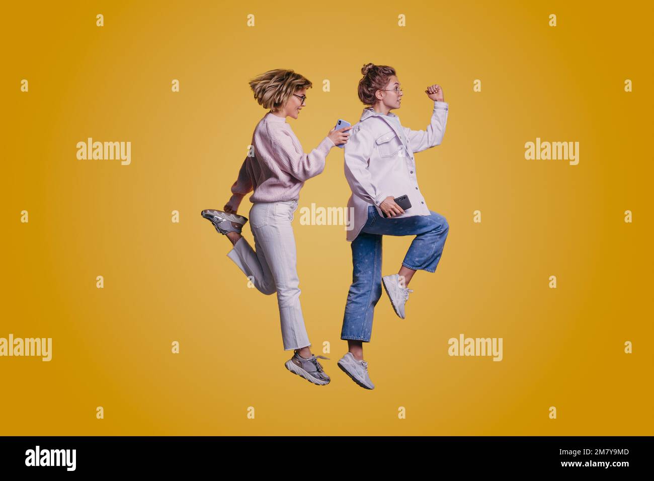 Ganzkörperansicht von zwei fröhlichen Frauen, die mit ihrem Telefon isoliert auf gelbem Hintergrund springen Stockfoto
