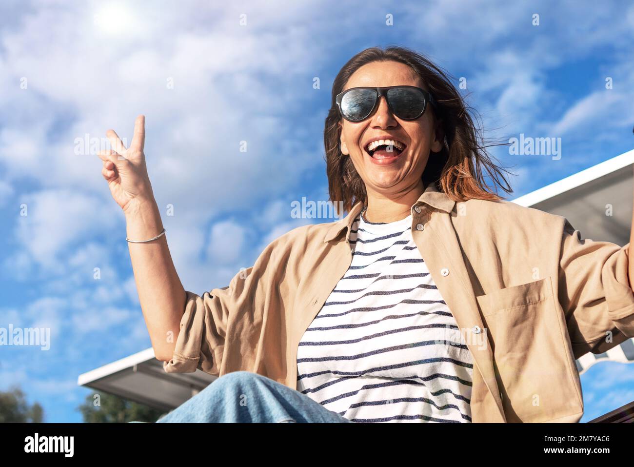 Eine glückliche, lächelnde Erwachsene Frau mit Sonnenbrille, die mit ihren Fingern im Freien gegen den blauen Himmel ein Siegessymbol gestalte. Stockfoto