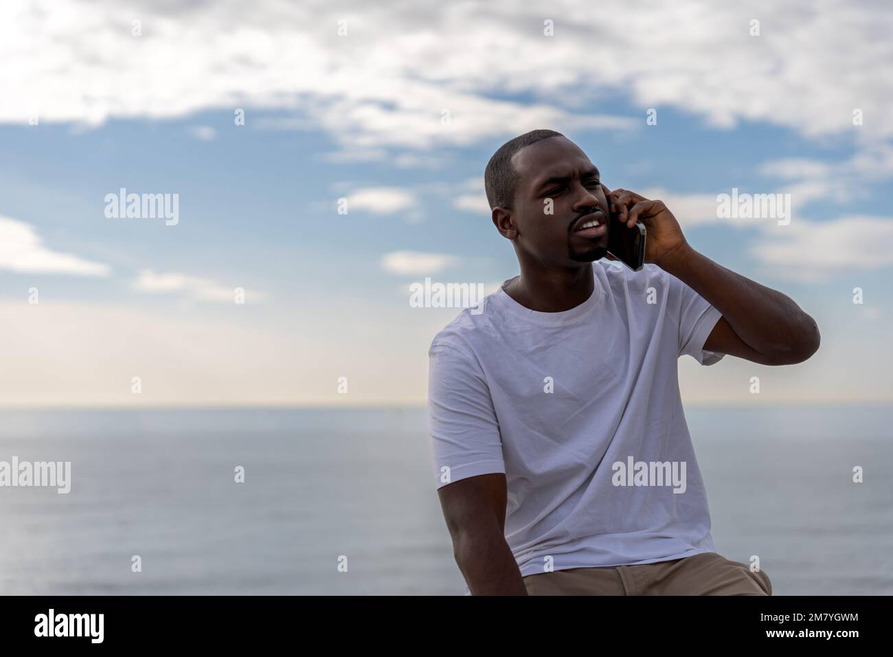 Ein seriöser afroamerikanischer Mann in lässiger Kleidung, der mit dem Handy spricht, während er unter dem Himmel gegen das Meer sitzt Stockfoto
